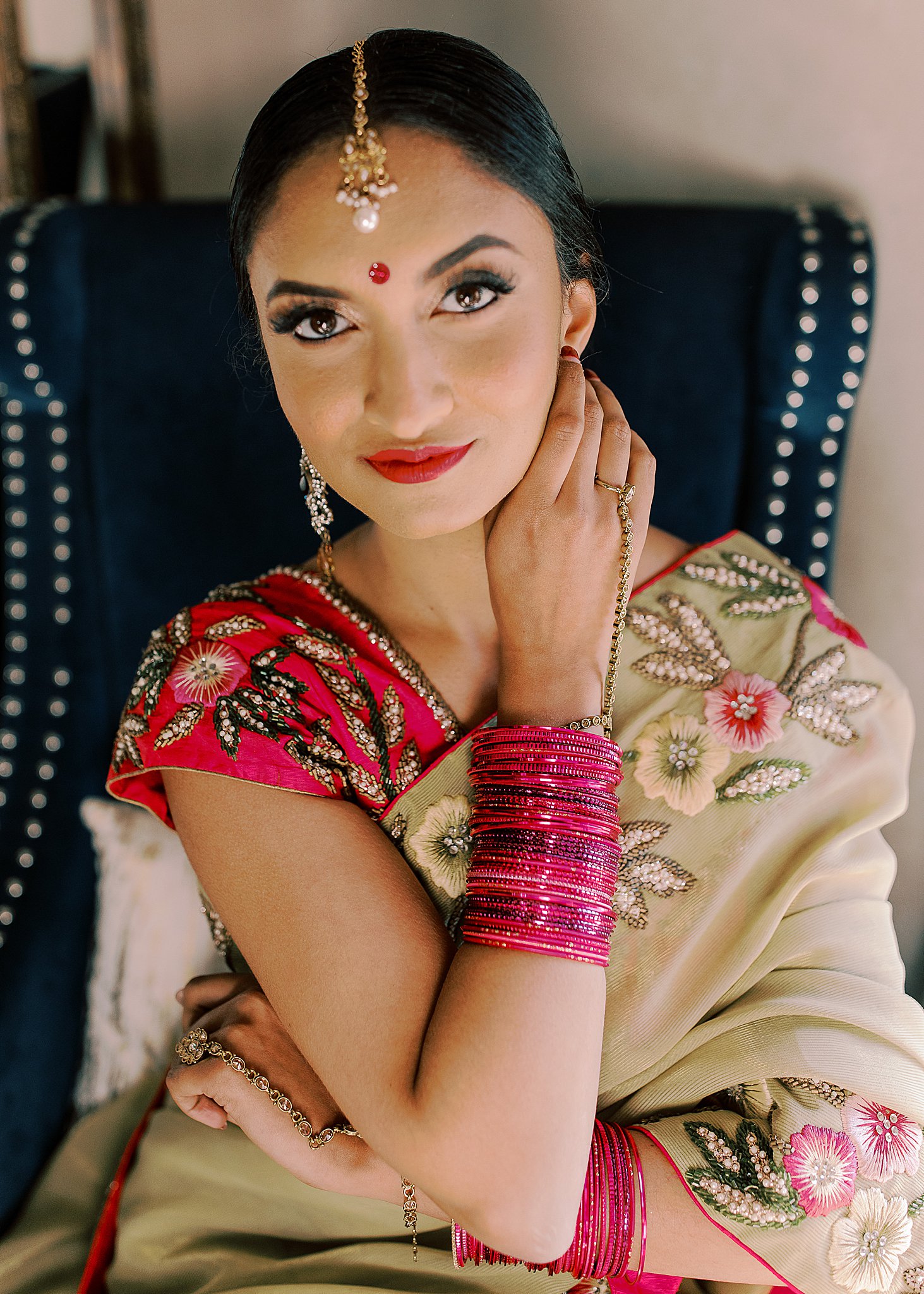 Beautiful Indian bride wearing hot pink wedding lehenga and bangles in blue velvet chair